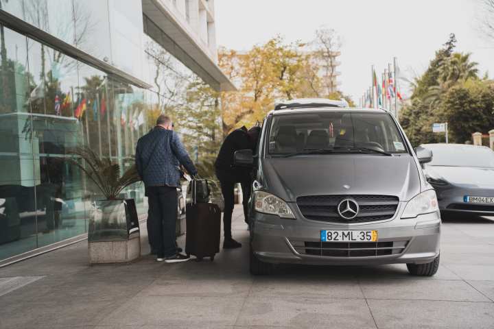 Two people loading luggage into a silver Mercedes van near a glass building entrance.