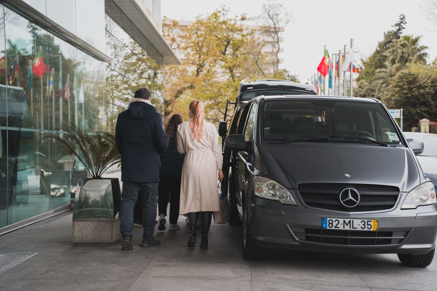 Three people walking past a parked gray Mercedes van near a glass building with international flags.