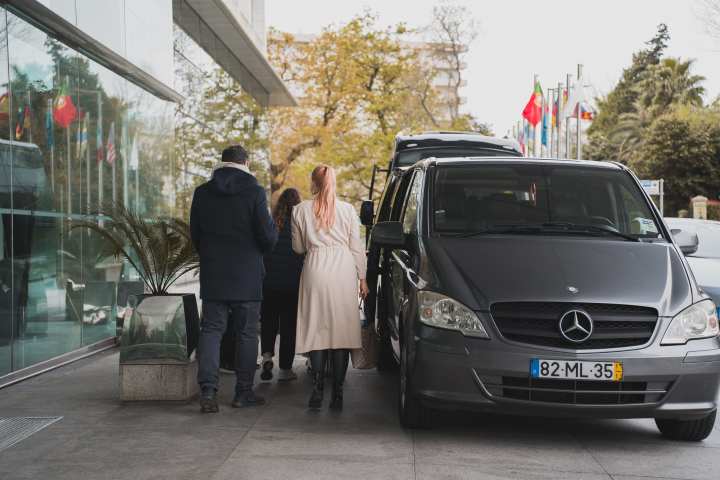 Three people walking past a parked gray Mercedes van near a glass building with international flags.