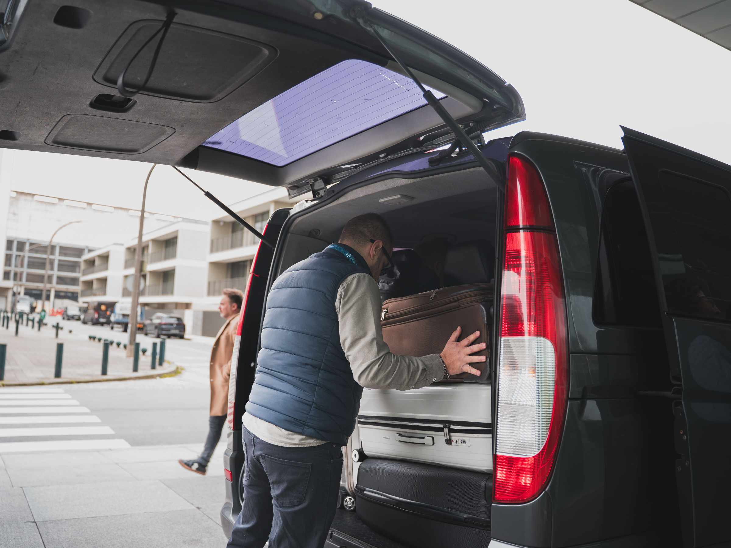 Man loads suitcase into the back of a minivan on a city street.