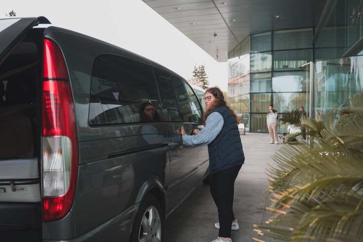 Person unloading luggage from a gray van in front of a modern building.
