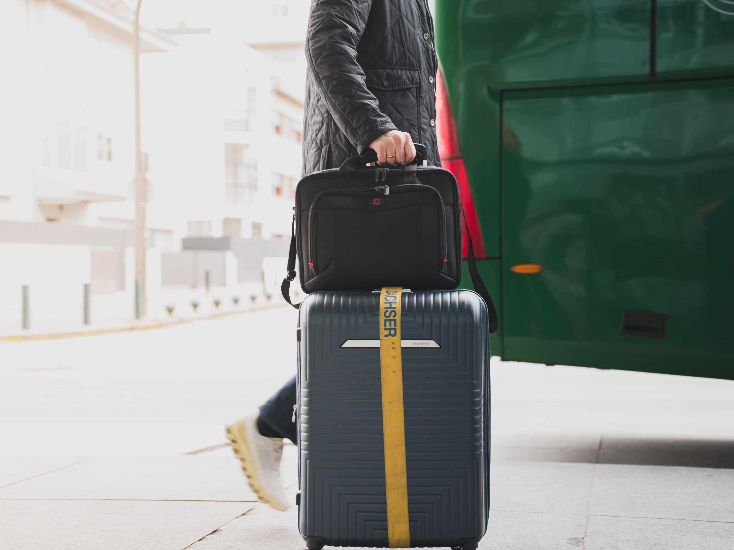 Person with suitcase and briefcase near a green bus.