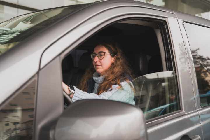 Woman with glasses driving a gray car, focused and wearing a scarf.