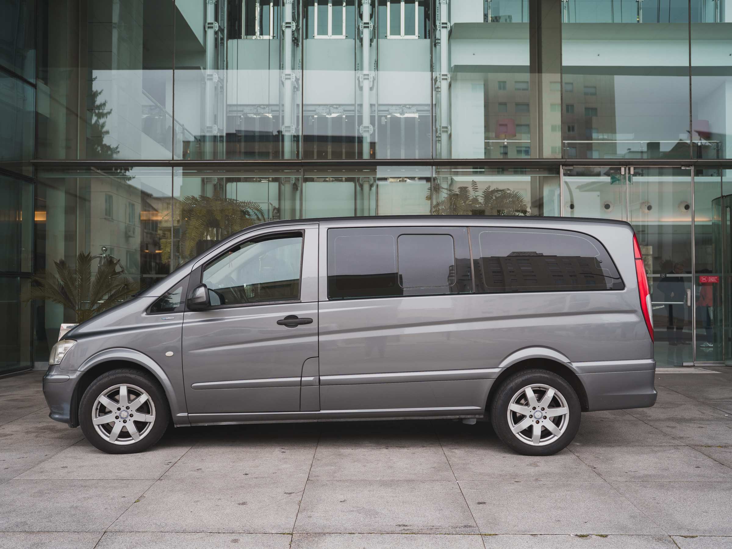 Side view of a grey van parked in front of a modern glass building.