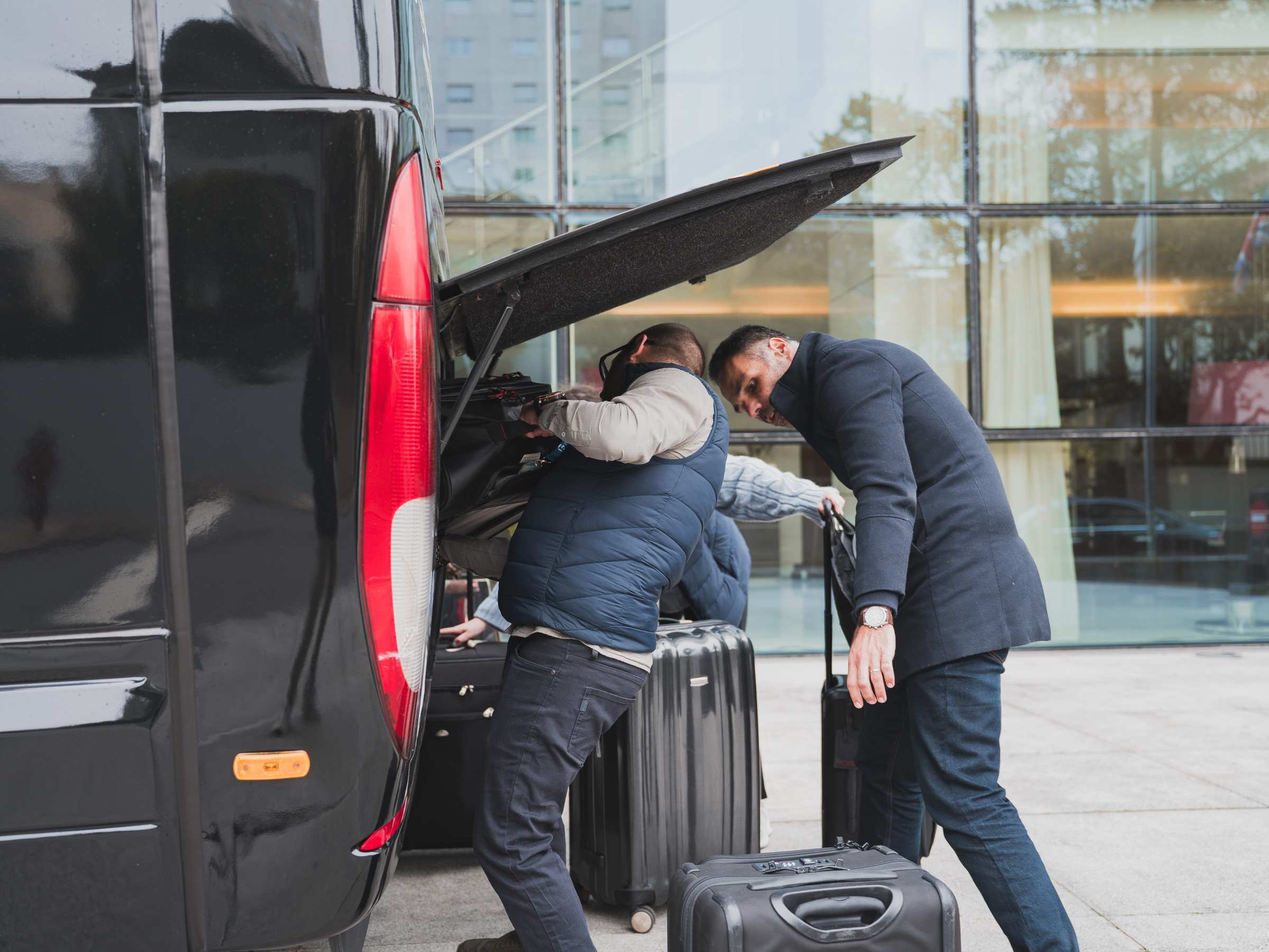 People loading luggage into the back of a black van outside a building.