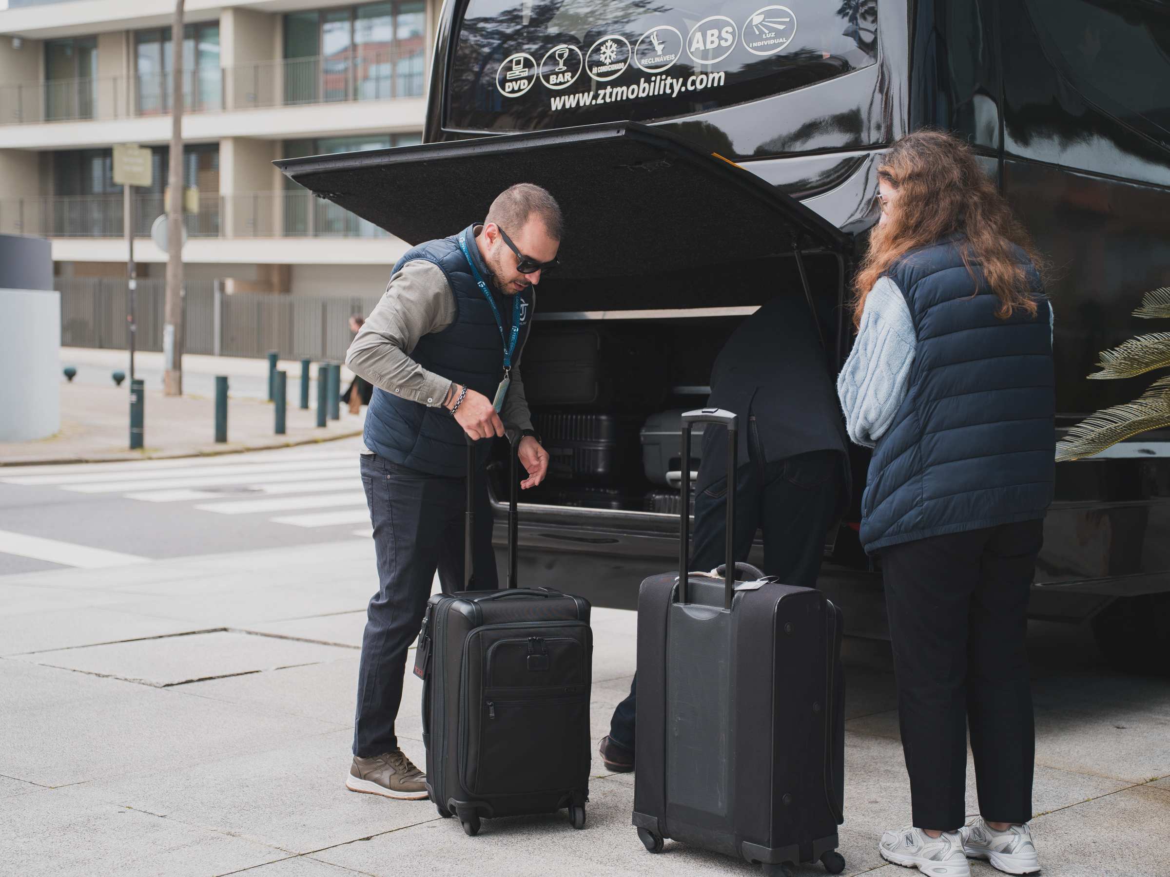 Two people loading suitcases into a van's open trunk on a city street.
