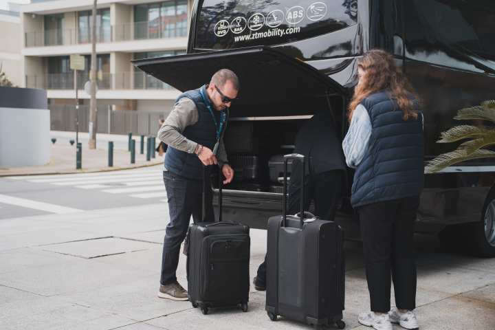 Two people loading suitcases into a van's open trunk on a city street.