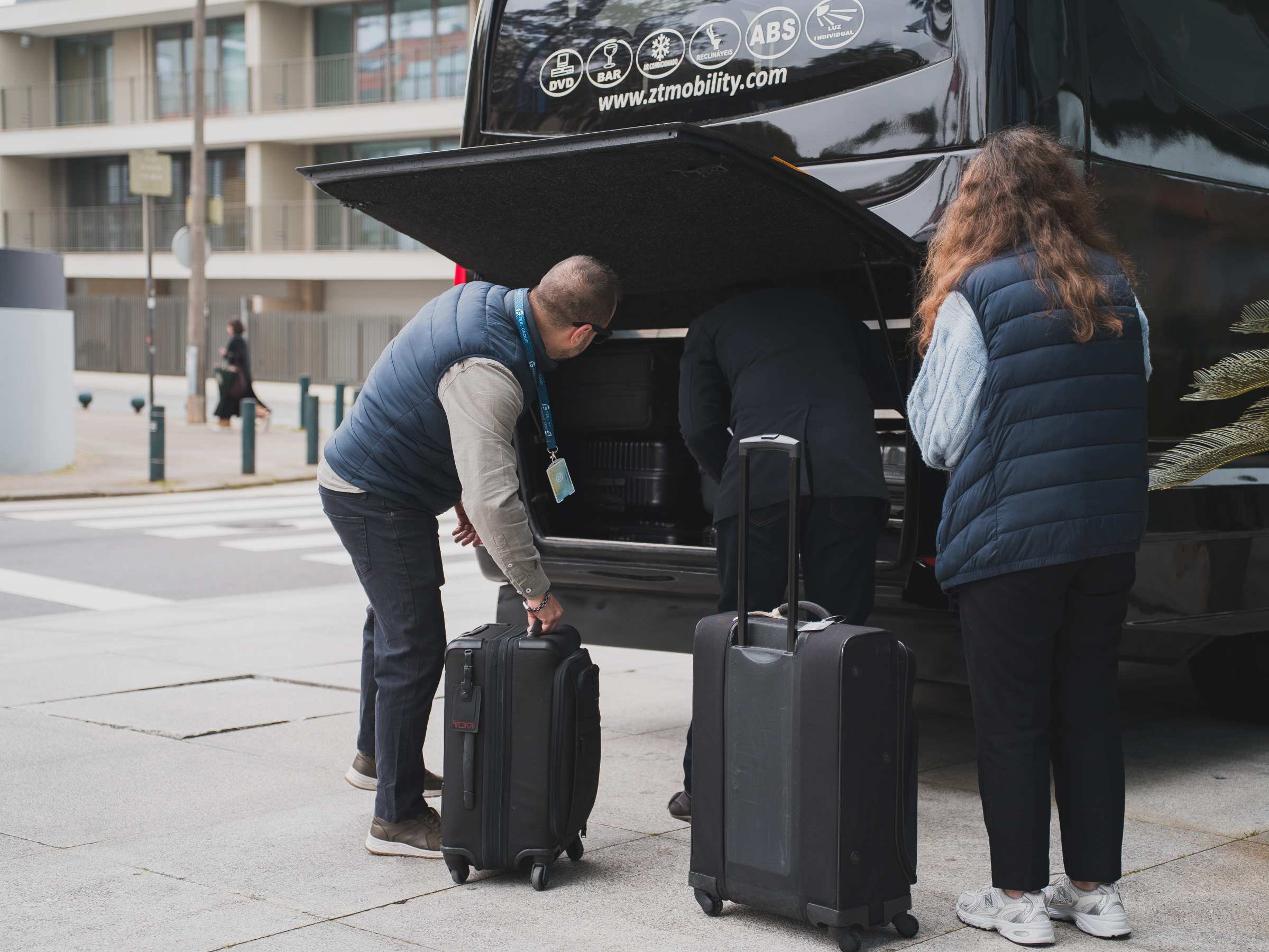 Three people loading luggage into a black van on a city street.