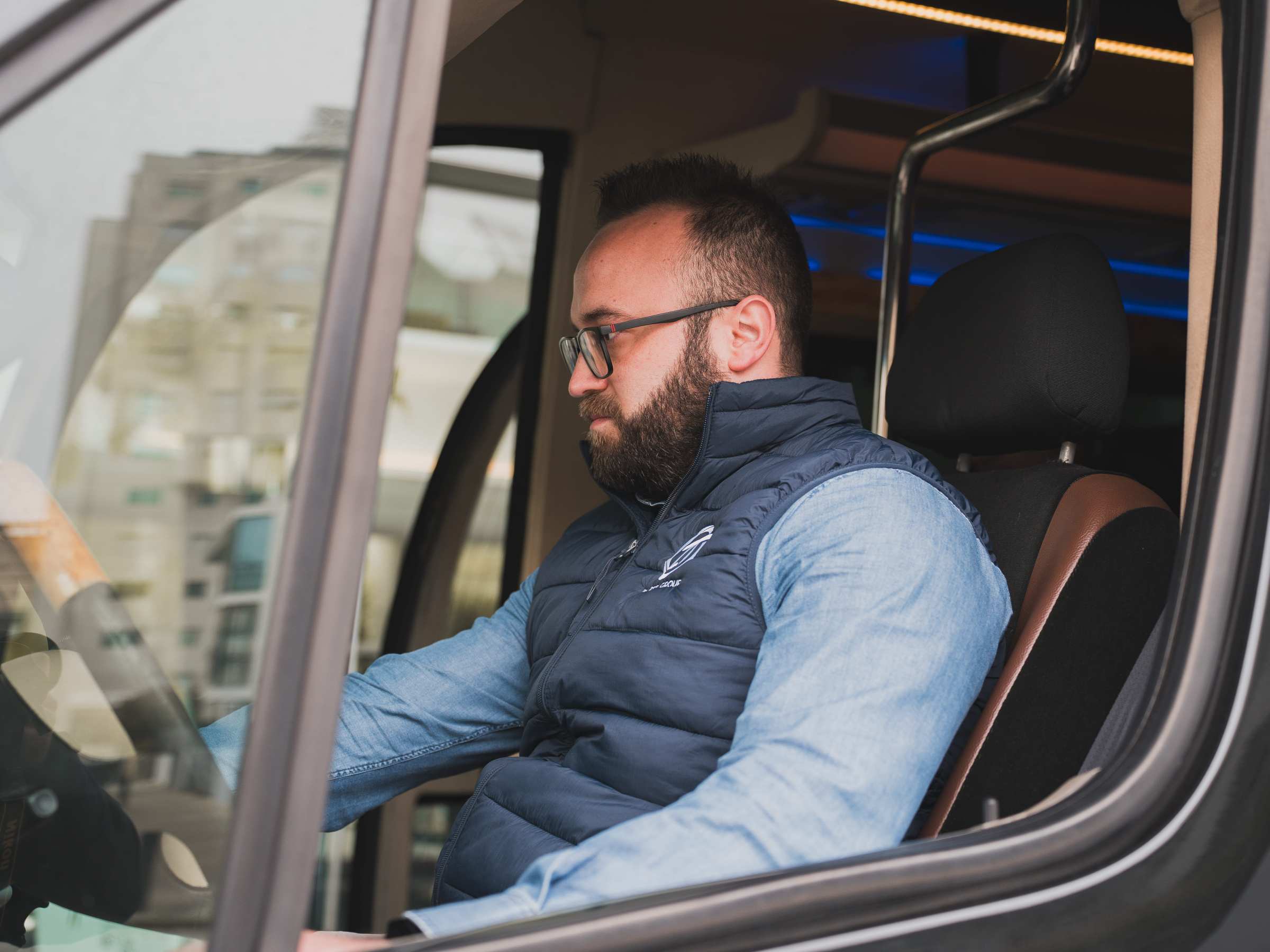 Man wearing glasses and blue vest sits in van driver's seat, looking forward.