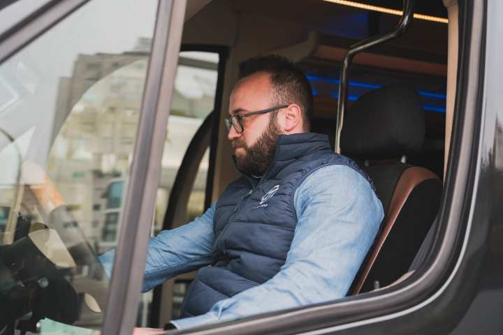 Man wearing glasses and blue vest sits in van driver's seat, looking forward.
