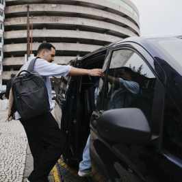 Man with backpack opening black car door on city street.