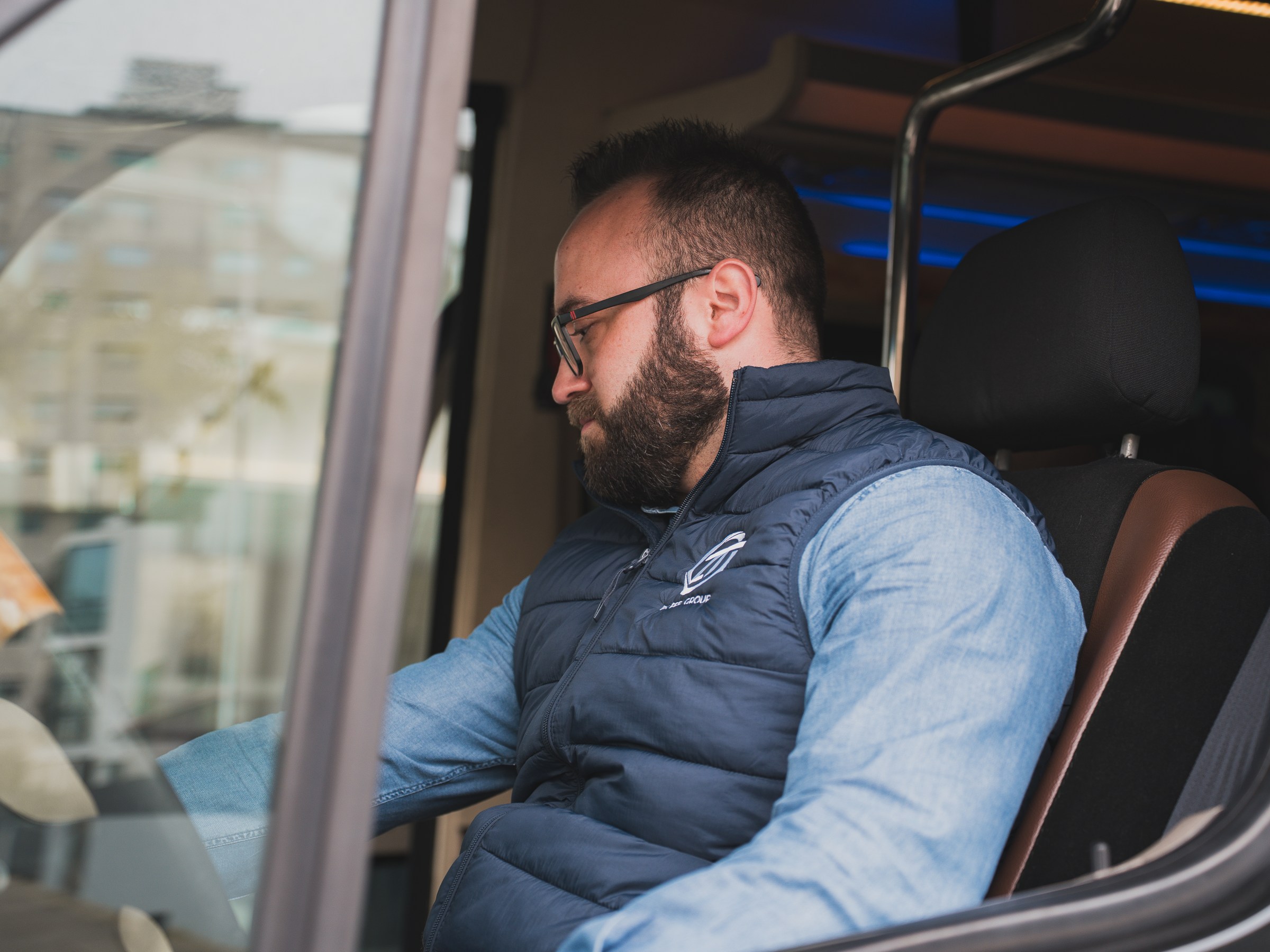 Man in blue vest seated inside vehicle, looking down.