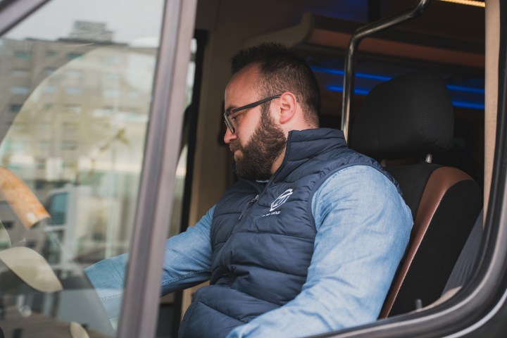 Man in blue vest seated inside vehicle, looking down.